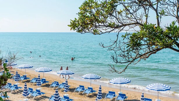 En strand med rader av blå solstolar och randiga parasoller, några personer i vattnet och en fri utsikt över havet.
