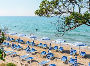 Una playa con hileras de sillas azules y sombrillas a rayas, algunas personas en el agua, y una vista despejada del mar.