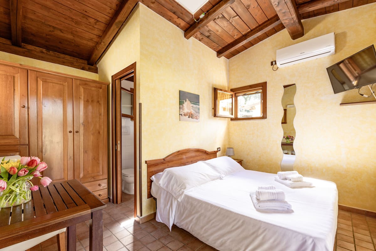 A bedroom with a wooden ceiling, a bed with white linens, a wooden table with a vase of flowers, and a bathroom visible through an open door.