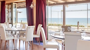 En restaurang med vita stolar och bord, röda draperier och utsikt över havet.