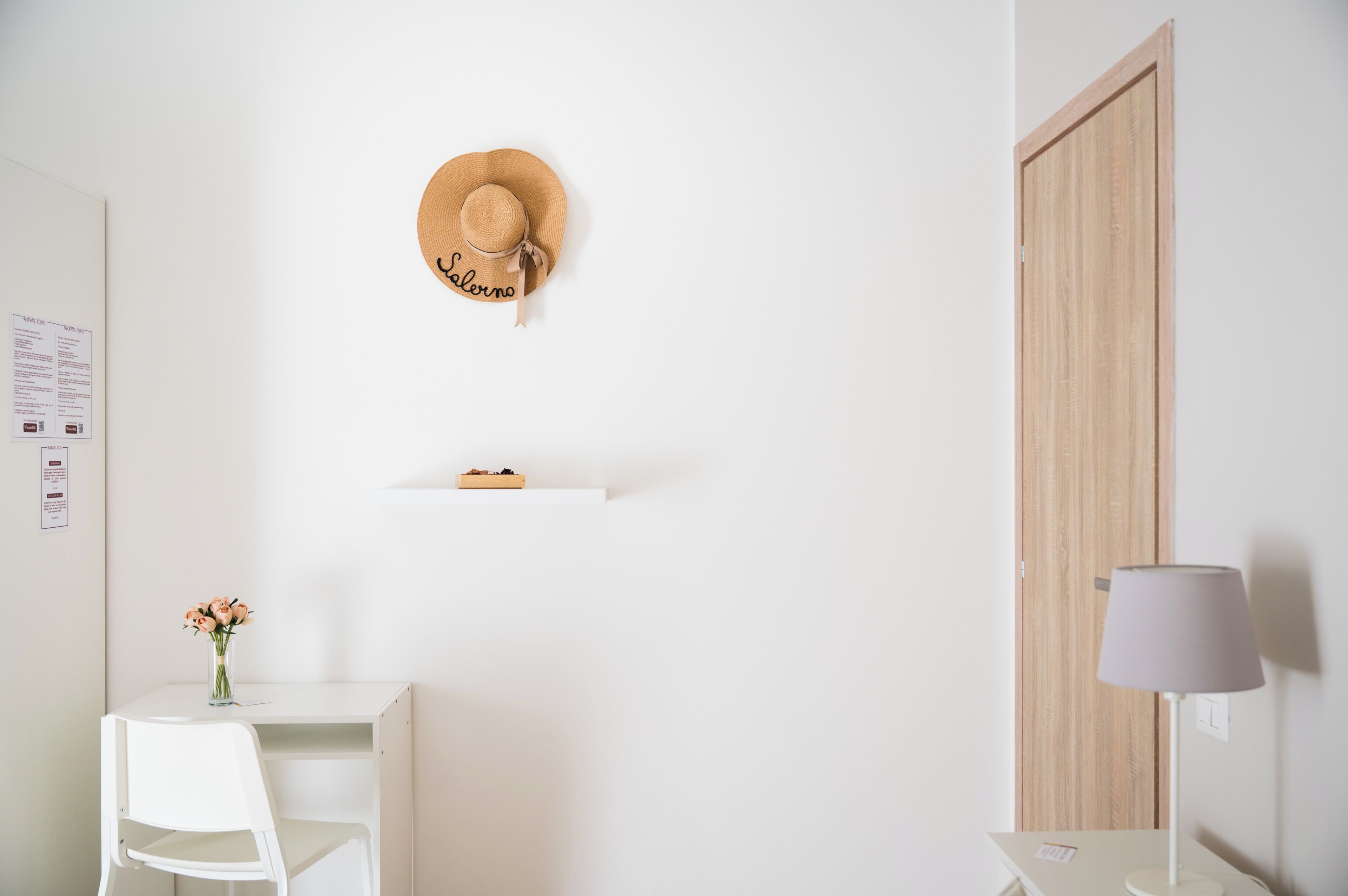 Habitación minimalista con un escritorio y una silla blancos, una puerta de madera, un sombrero colgado en la pared, un estante pequeño con una caja y un jarrón con flores.