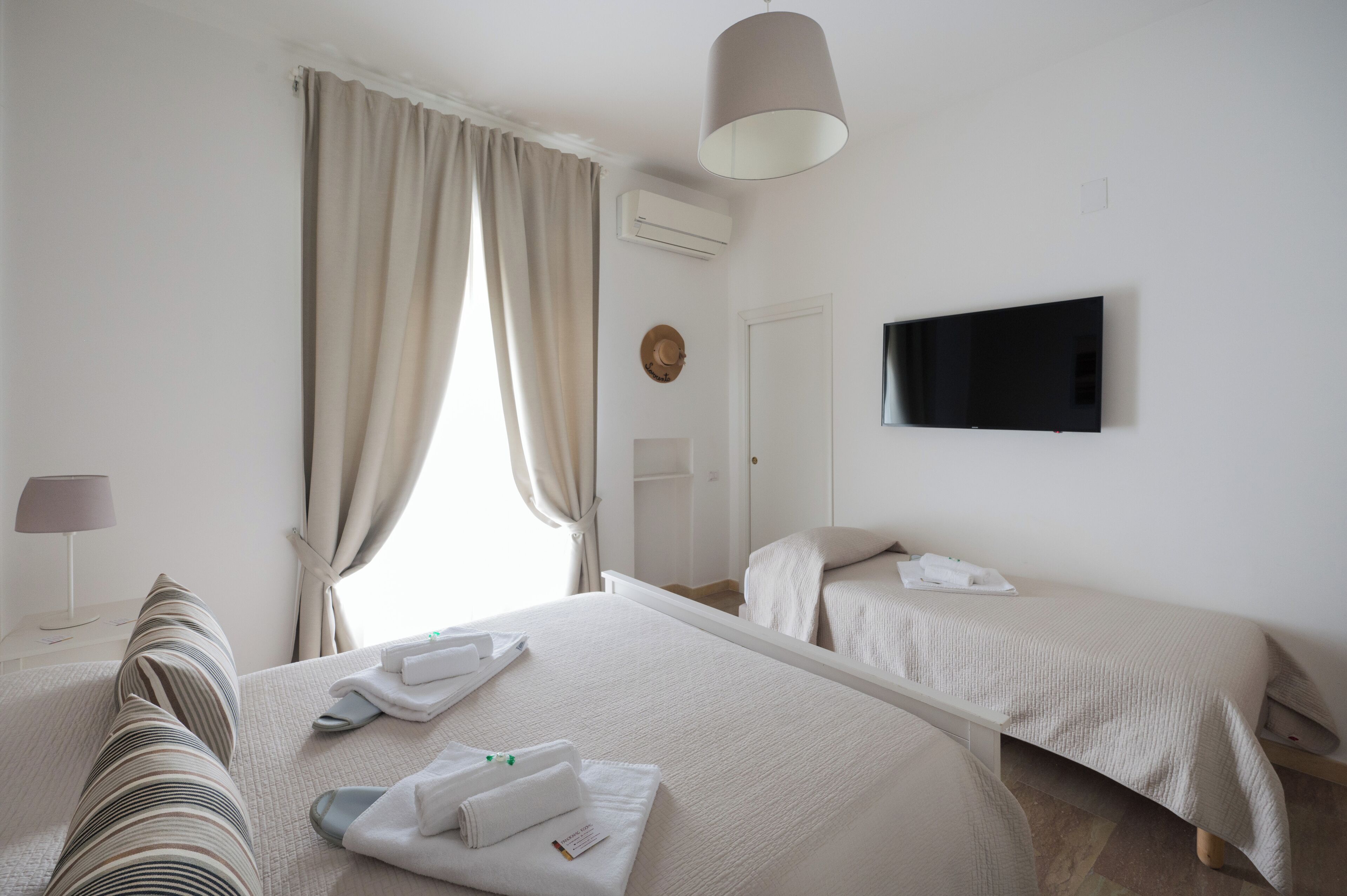 Habitación de hotel con una cama grande, dos camas más pequeñas, un televisor de pantalla plana, una lámpara y un ventanal con cortinas.