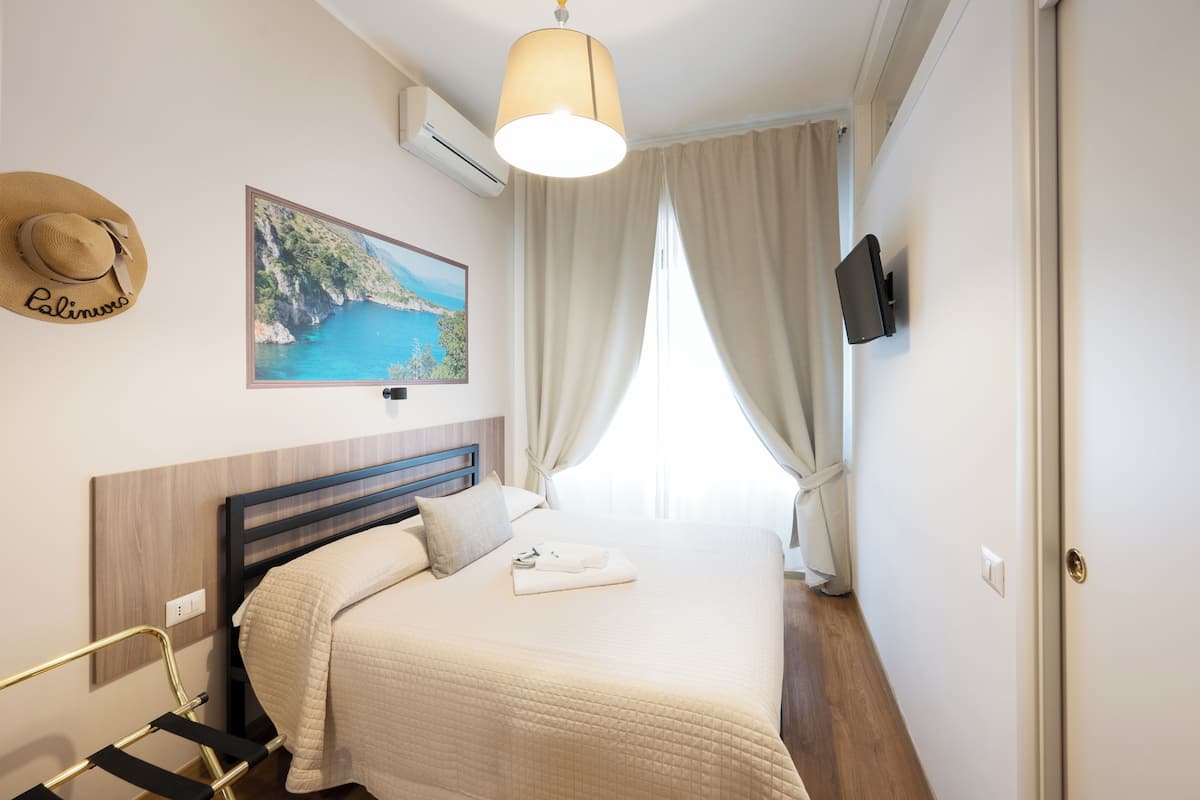 Une chambre d’hôtel avec un lit, un tableau représentant un paysage côtier, un chapeau accroché au mur et un téléviseur fixé au mur.