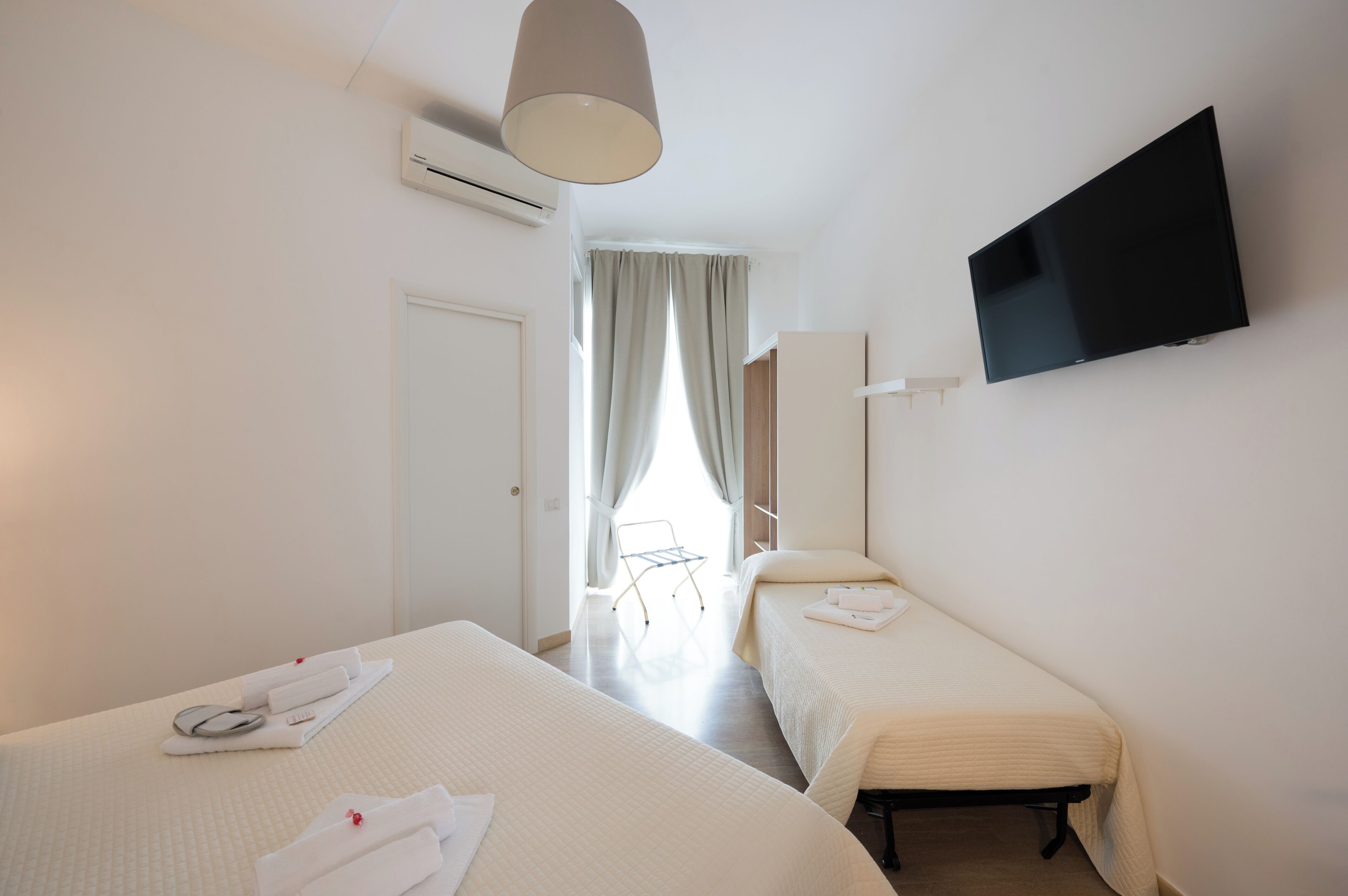 A hotel room with two beds, a TV, an air conditioning unit, and a window with curtains.