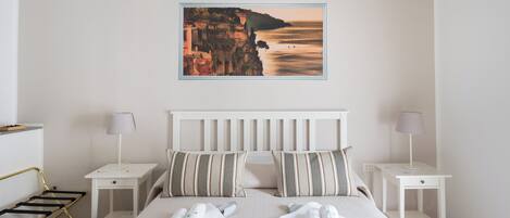 A hotel room with a bed, two bedside tables, a bench, and a painting of a coastal scene.