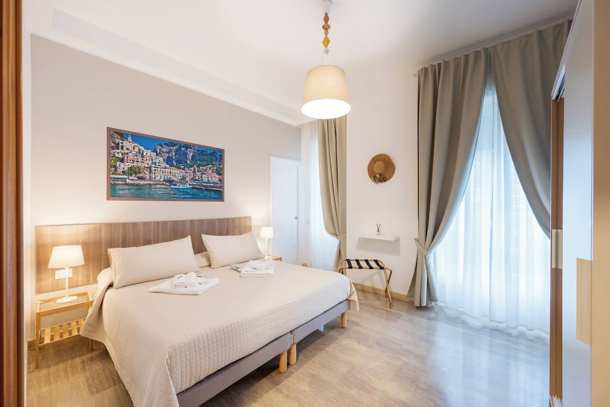 Une chambre d’hôtel avec un grand lit, une table de chevet avec une lampe, un tableau représentant une ville côtière et une vue sur la mer à travers de fines rideaux.