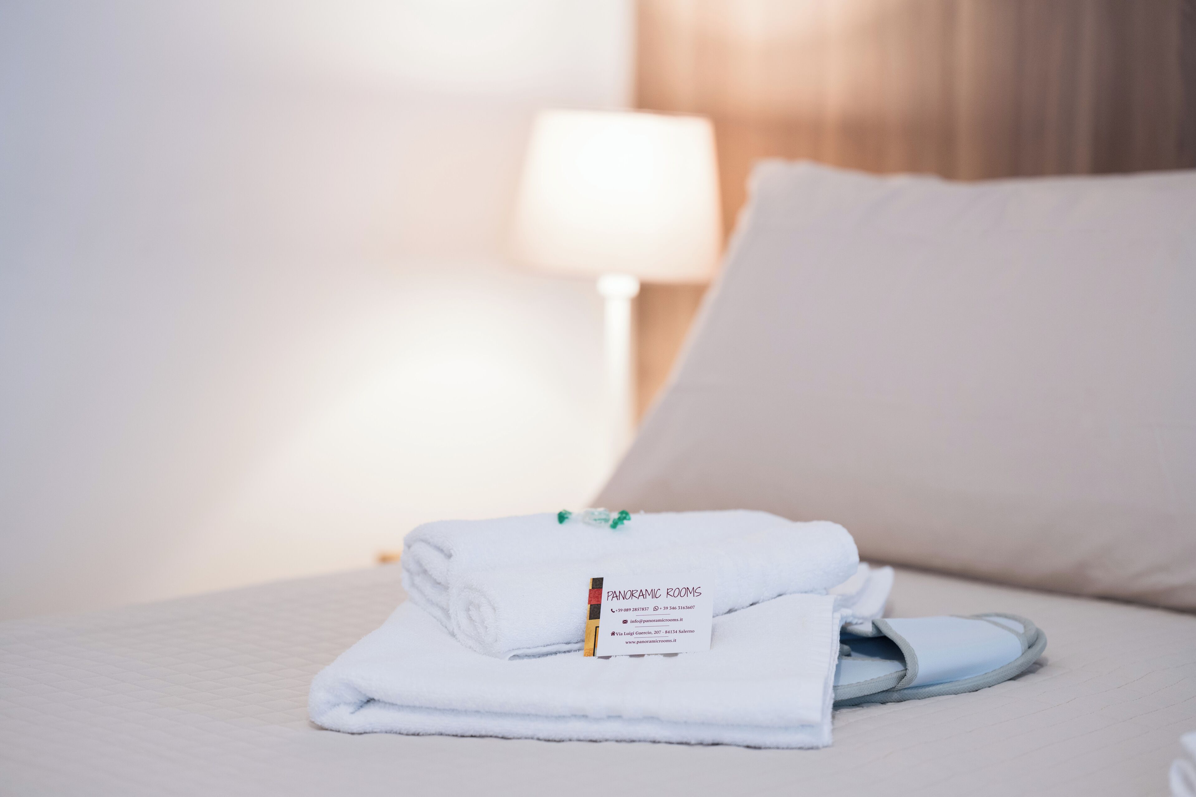 Une serviette blanche soigneusement pliée et une paire de chaussons bleu clair sont posées sur un lit avec un oreiller blanc et une lampe en arrière-plan.