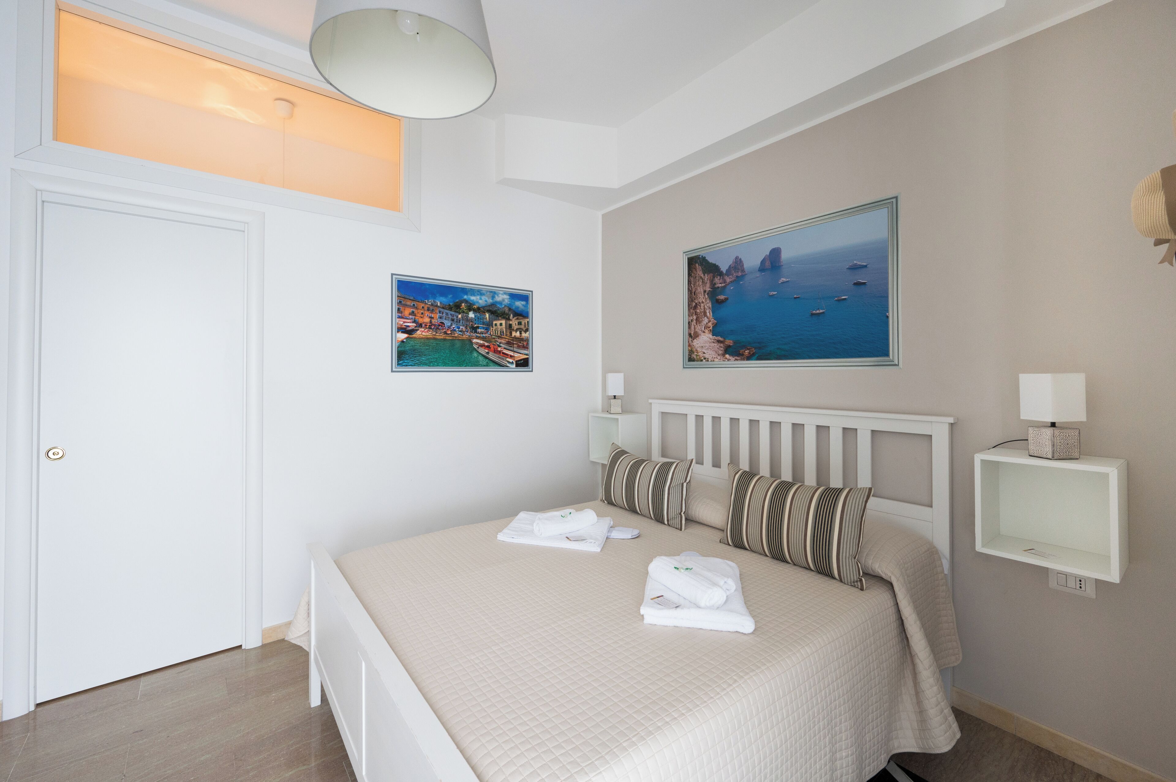 A neatly arranged bedroom with a bed, two paintings, a bedside table with a lamp, and a door.
