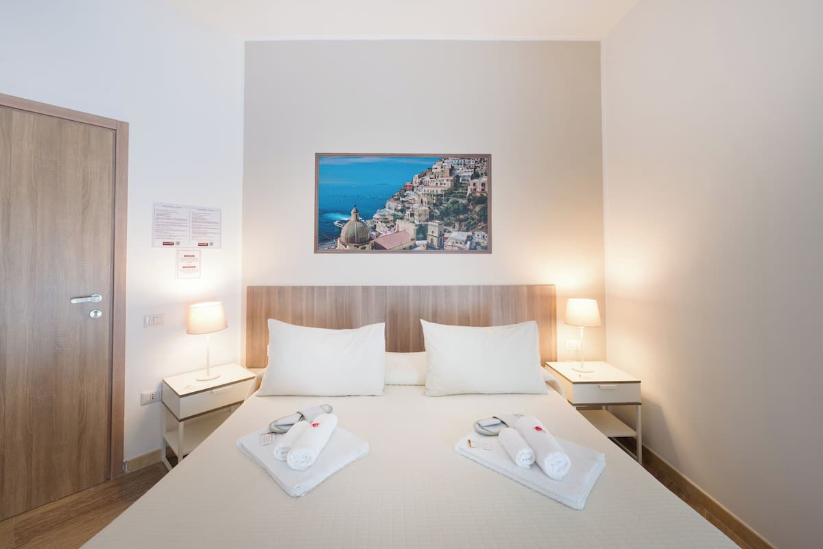 Une chambre d’hôtel avec un lit, deux tables de chevet, une porte en bois, un tableau accroché au mur et un petit panneau.