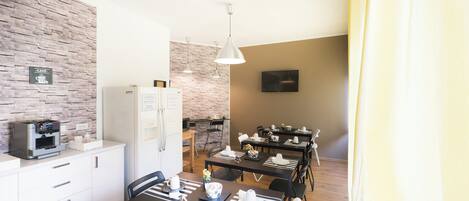 Una cocina moderna con cafetera, armarios blancos y una pared de ladrillo decorativa. Hay un comedor con mesas dispuestas para cuatro personas, un refrigerador y un televisor de pantalla plana montado en la pared.