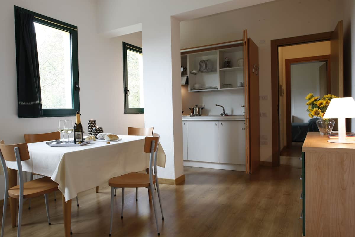Un coin repas avec une table dressée pour deux personnes, une cuisine avec des placards ouverts et une vue sur l’extérieur par une fenêtre.