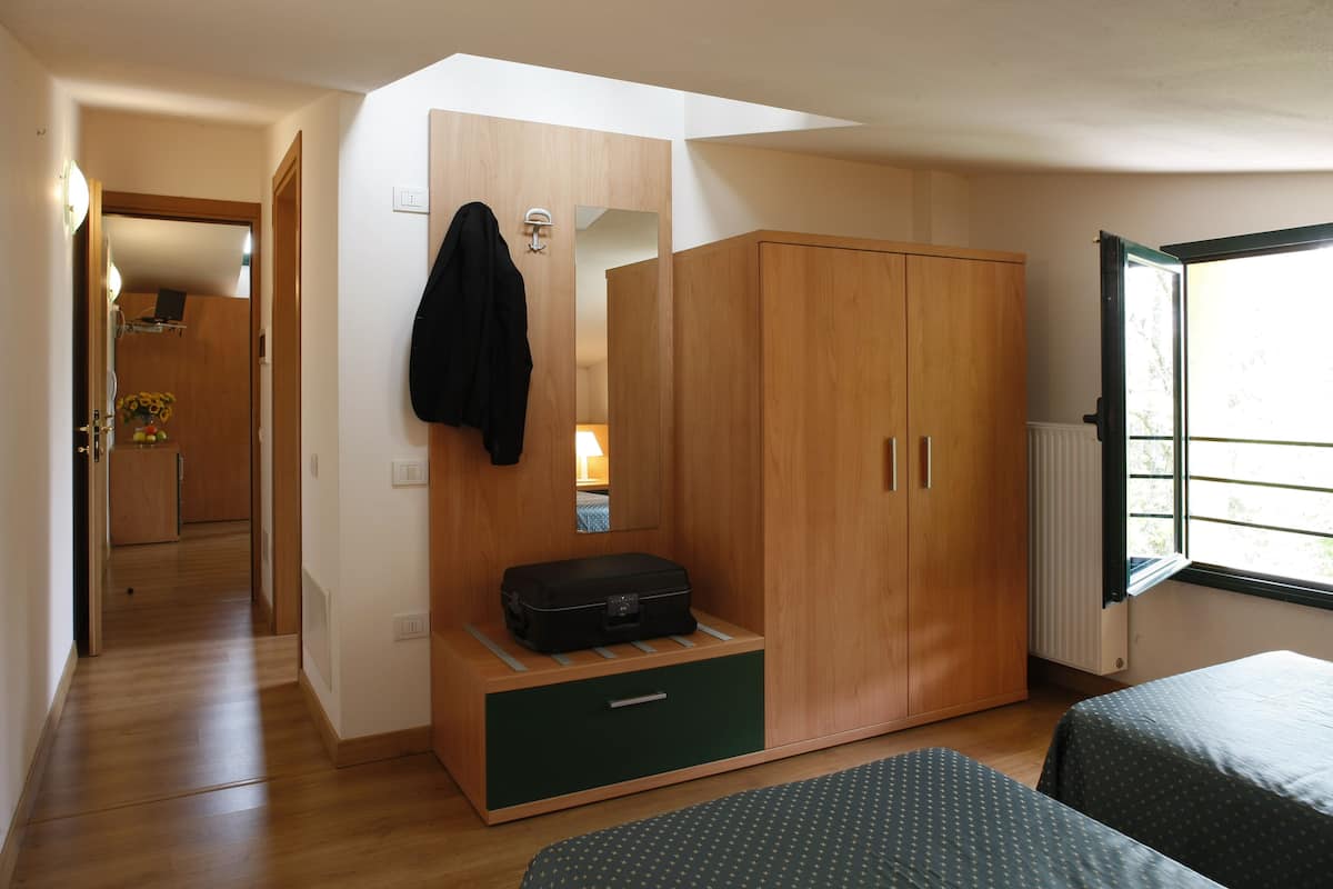 Une chambre à coucher avec une armoire en bois, un miroir, une valise posée sur une étagère et une fenêtre donnant sur un paysage.