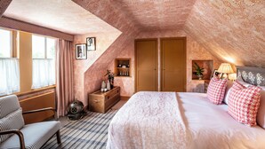 An attic bedroom with a sloped ceiling, a bed with patterned bedding, a chest of drawers, a vintage-style lamp, and a window with curtains. - Loch Lomond Arms Hotel (Alexandria)