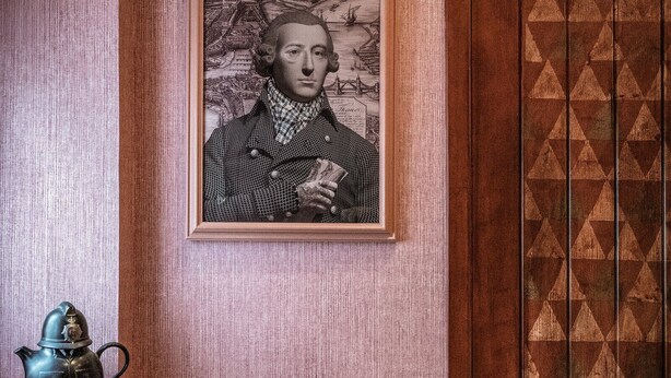 A room with a framed portrait, a wooden door with geometric patterns, a chair with a patterned cushion, and a small table with a teapot. - Loch Lomond Arms Hotel (Alexandria)