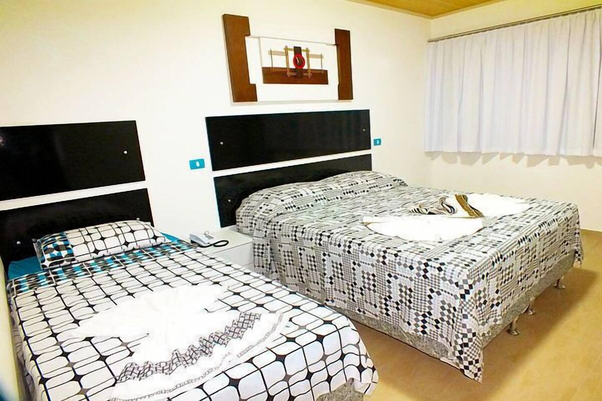Een hotelkamer met twee bedden, elk met een zwart hoofdeinde en een bedsprei met een patroon. Er is een raam met witte gordijnen en een klok aan de muur.