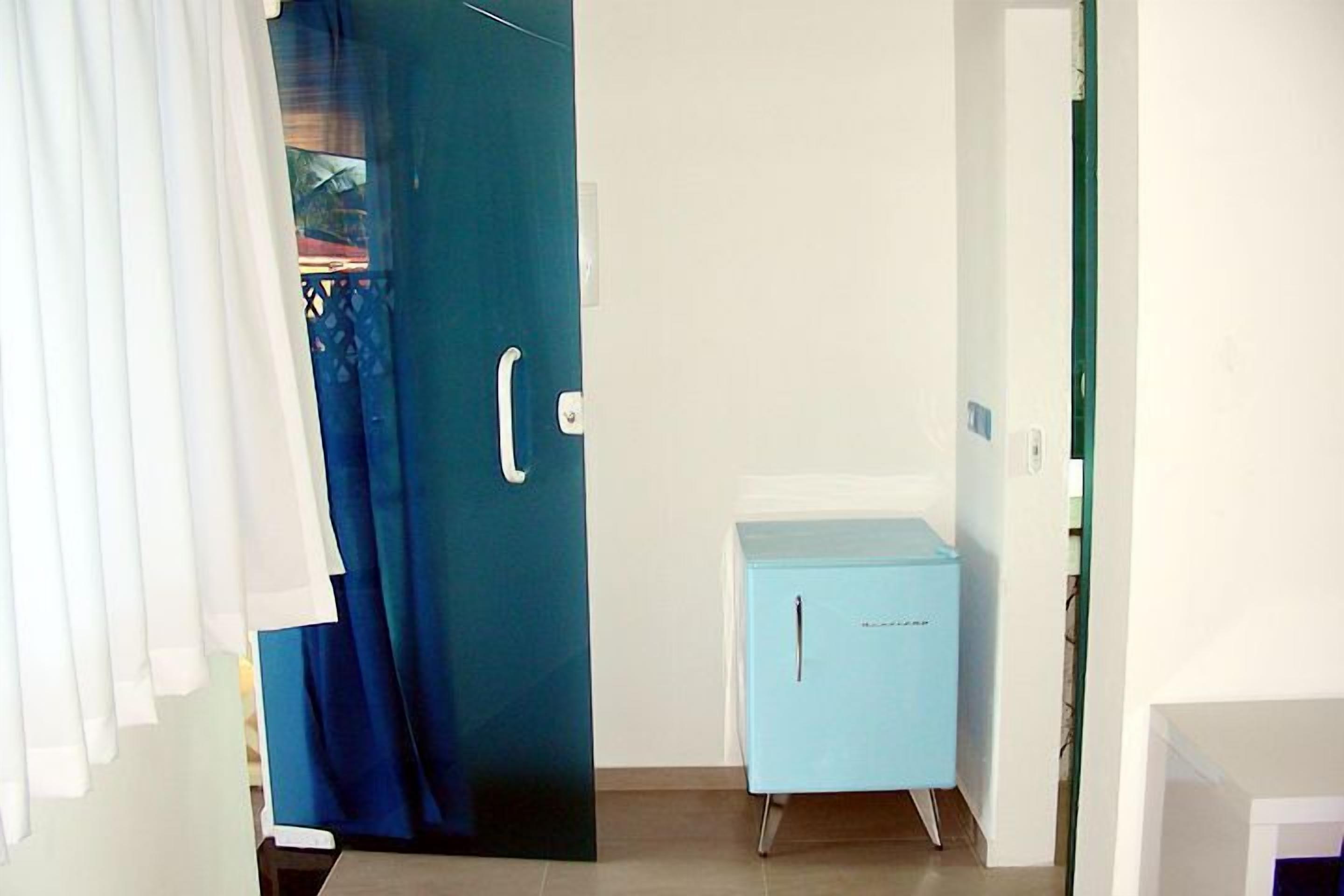Una habitación con una puerta cerrada, un pequeño armario azul y una cortina entreabierta.