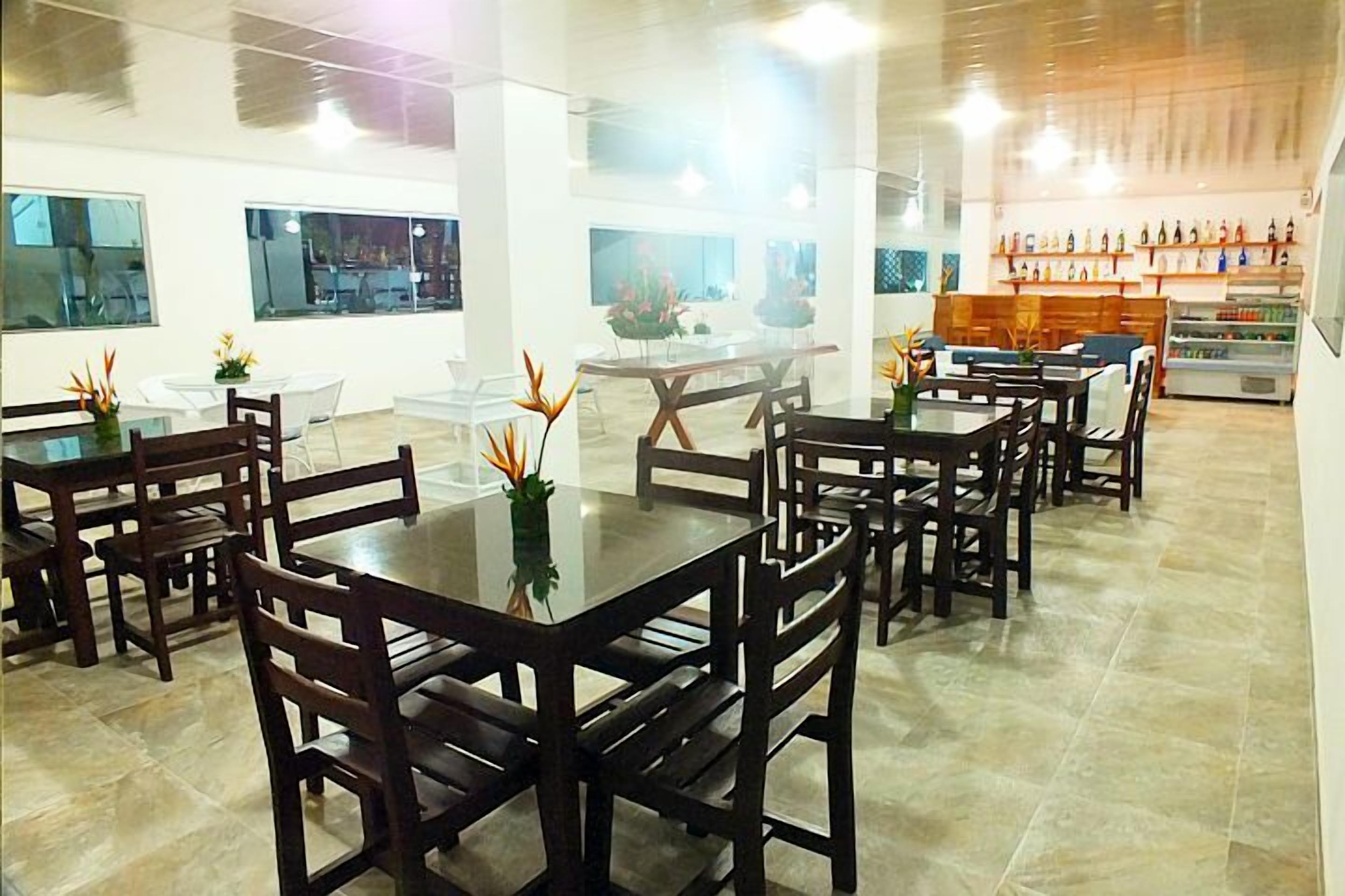 Uma ampla sala de jantar com várias mesas e cadeiras, grandes janelas e uma seleção de bebidas engarrafadas.