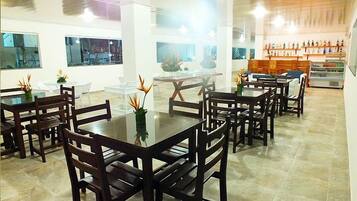 Uma ampla sala de jantar com várias mesas e cadeiras, grandes janelas e uma seleção de bebidas engarrafadas.