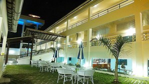 Une terrasse extérieure avec des chaises et des tables blanches, un palmier et un bâtiment doté de grandes fenêtres et d’un escalier.