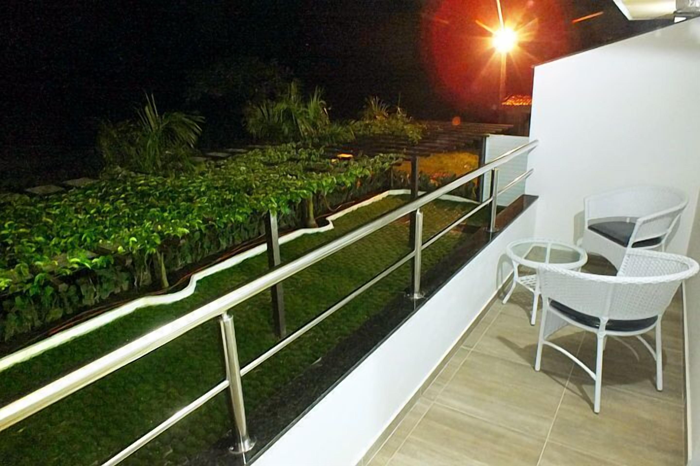 Un balcon agrémenté de chaises en osier blanc et d’une petite table, donnant sur un jardin bien entretenu où poussent différentes espèces de plantes, et bordé d’un sentier.