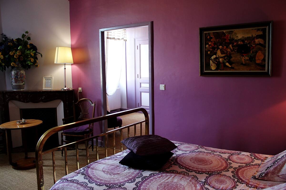 Une chambre à coucher avec un lit, une chaise, une table, une lampe, un tableau et un vase de fleurs.