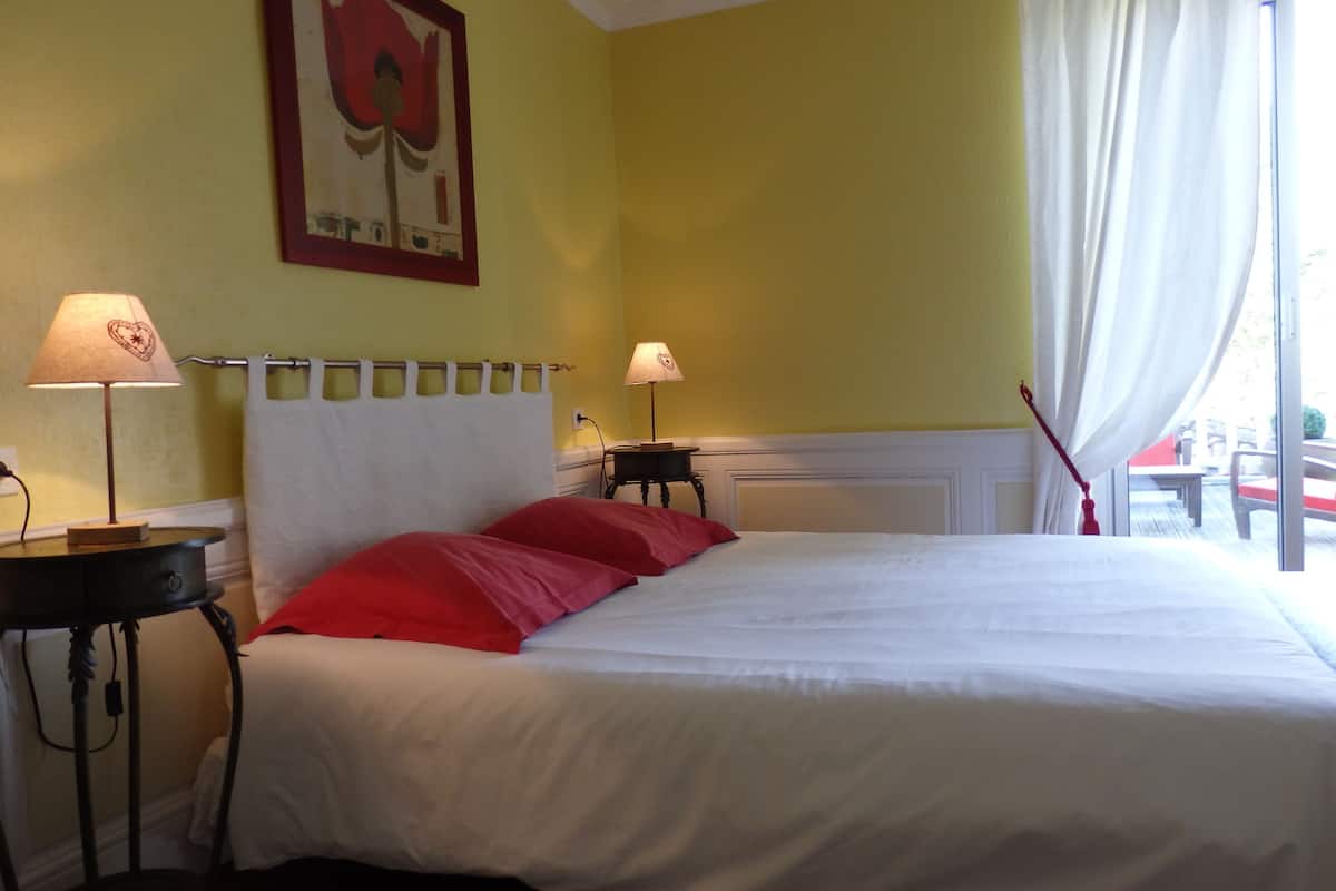 Une chambre d’hôtel avec un lit, deux tables de chevet, une commode, un tableau et une fenêtre avec des rideaux.