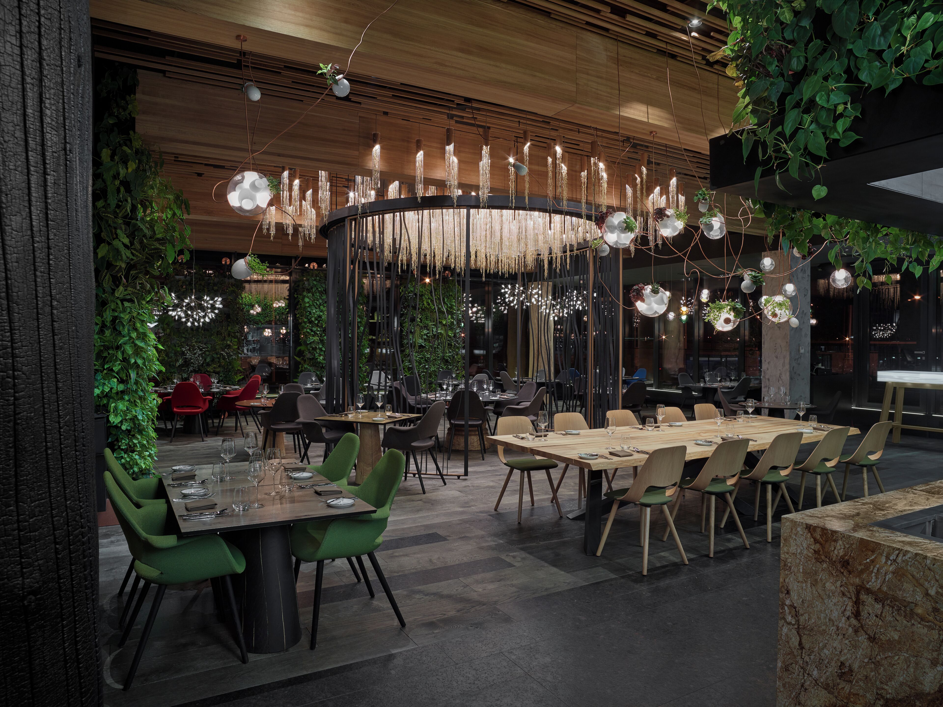 Een modern restaurant met houten tafels en stoelen, hangende lampen en groene planten.
