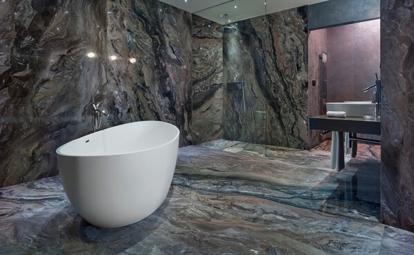 Un baño moderno con una bañera ovalada grande y blanca, una ducha con cerramiento de vidrio y una zona de lavabo en tonos oscuros.
