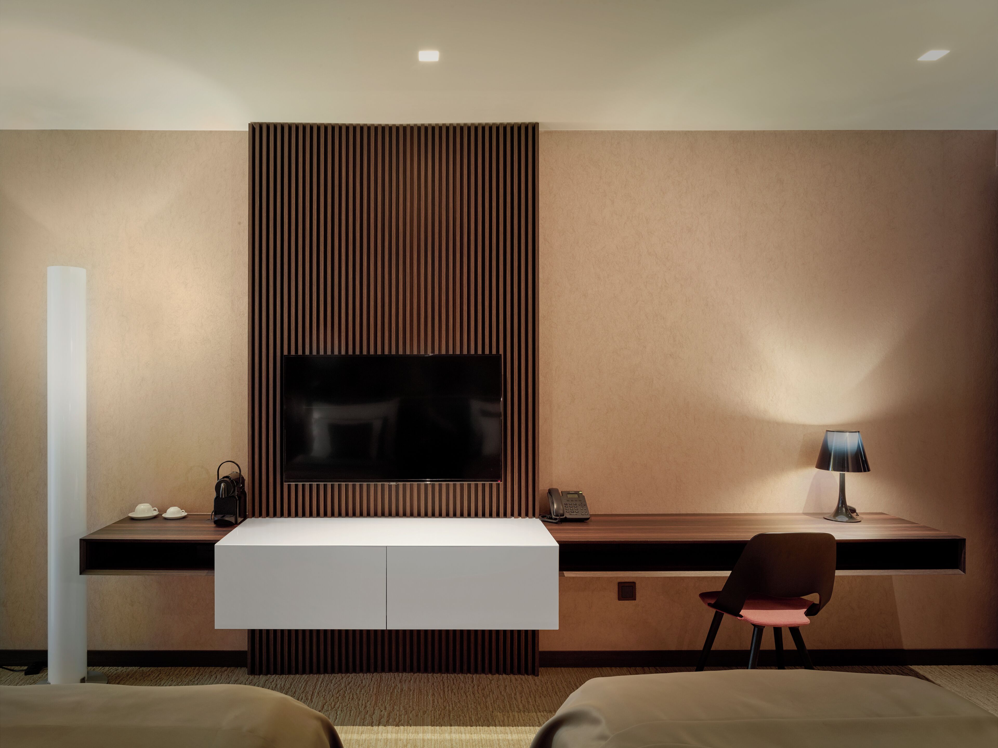Una habitación de hotel moderna con un televisor de pantalla plana grande montado en una pared de paneles de madera, un escritorio con lámpara, una silla y una cama con cabecera.