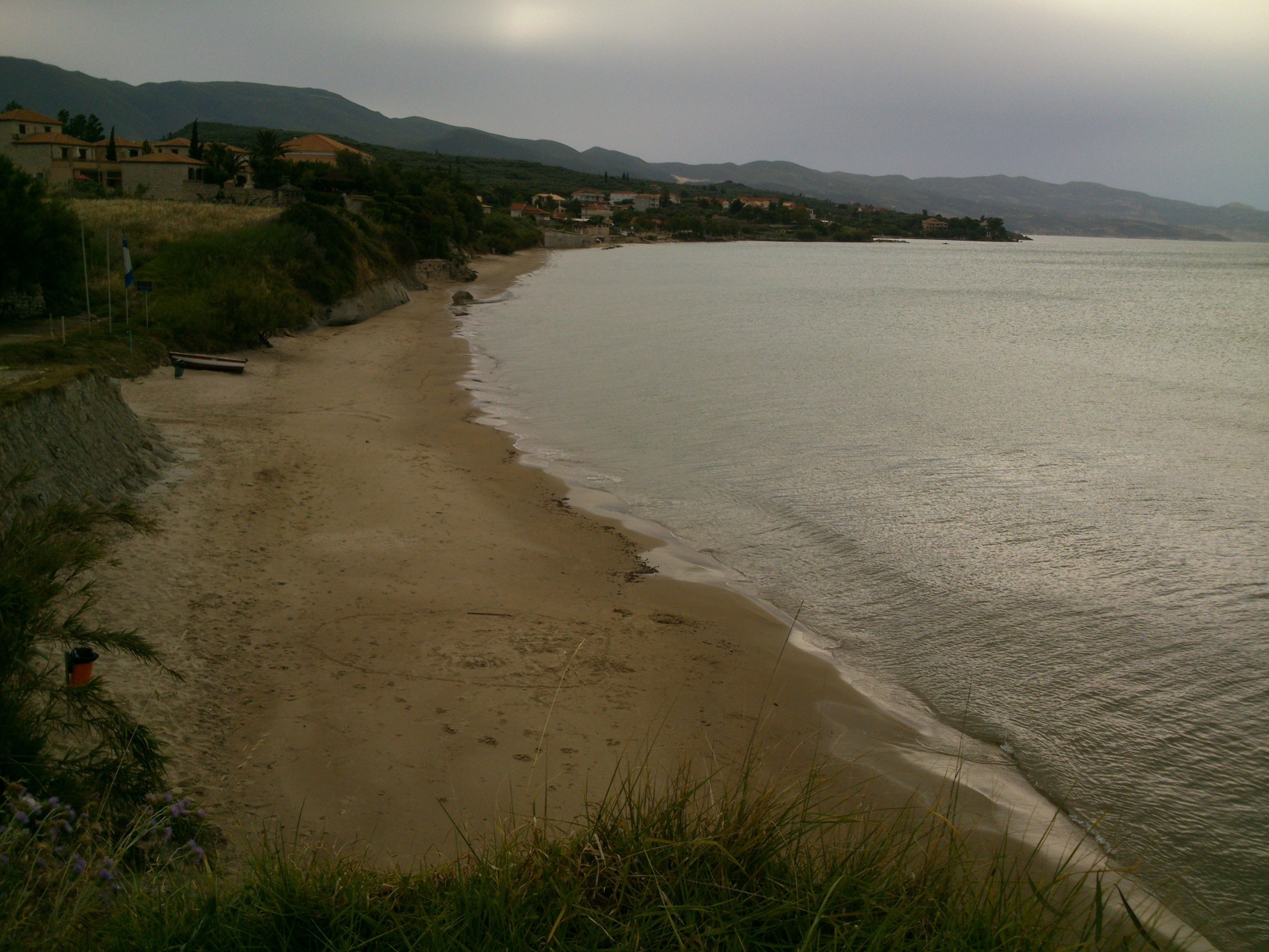 A beach with a sandy shore, a bench, and a view of houses and mountains in the background.