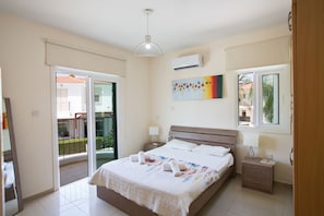 A hotel bedroom with a large bed, a nightstand, a lamp, an air conditioner, and a sliding glass door to the balcony. - Shaye Frontline Villa (Paralimni)
