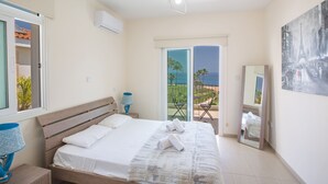 A bedroom with a bed, a nightstand, a lamp, a balcony with a view, and a painting on the wall. - Shaye Frontline Villa (Paralimni)