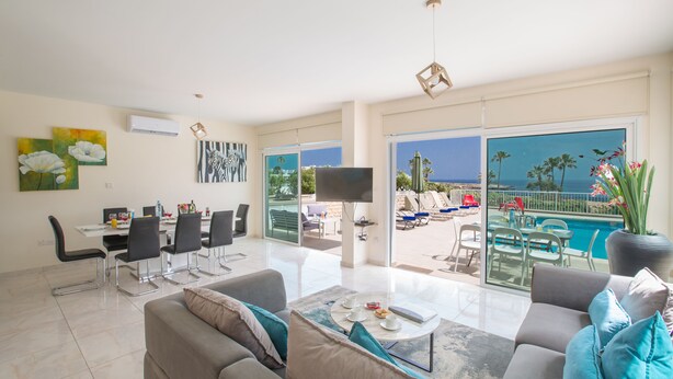 A spacious living room with a sofa, coffee table, and dining area. A large TV is mounted on the wall, and there is a balcony with a pool view. - Shaye Frontline Villa (Paralimni)