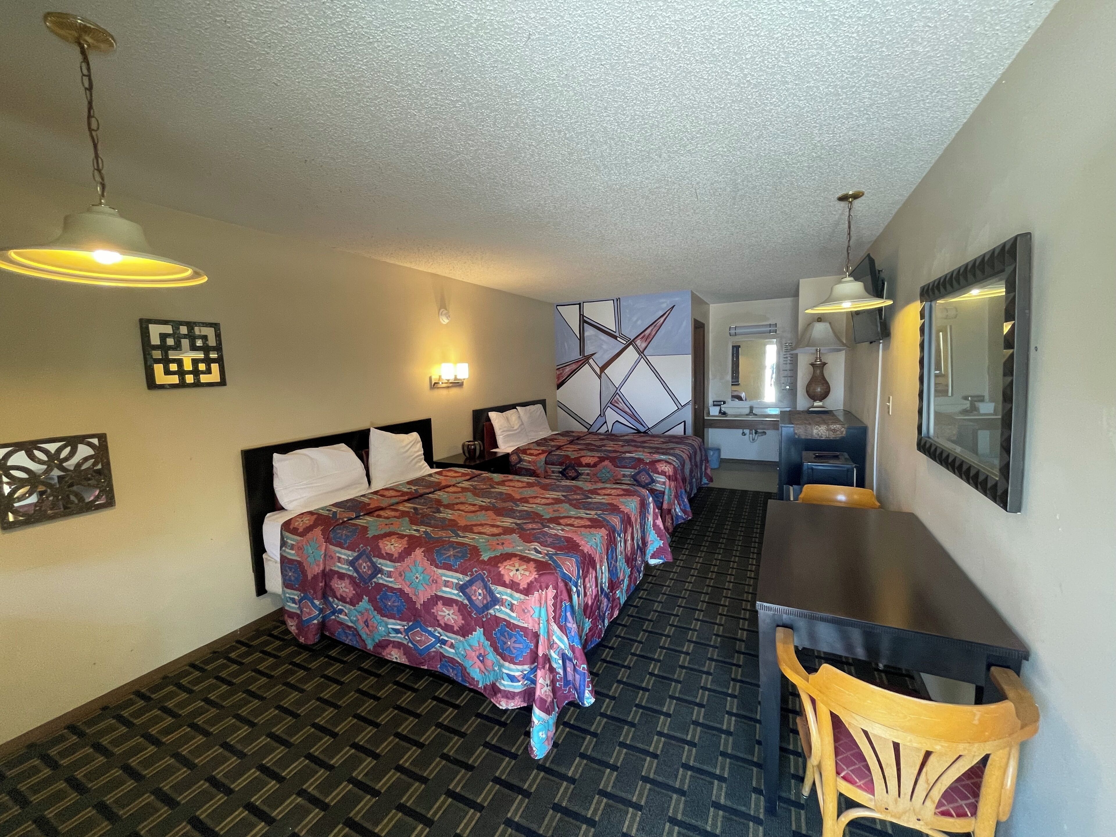 A hotel room with two beds, a desk, and a chair. There is a mirror, a wall clock, and a decorative wall piece.