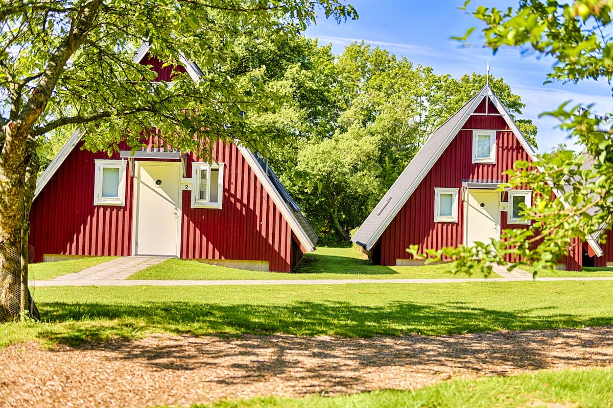 Two red wooden cabins with white trim, each featuring a door and a window, set in a grassy area with trees in the background.