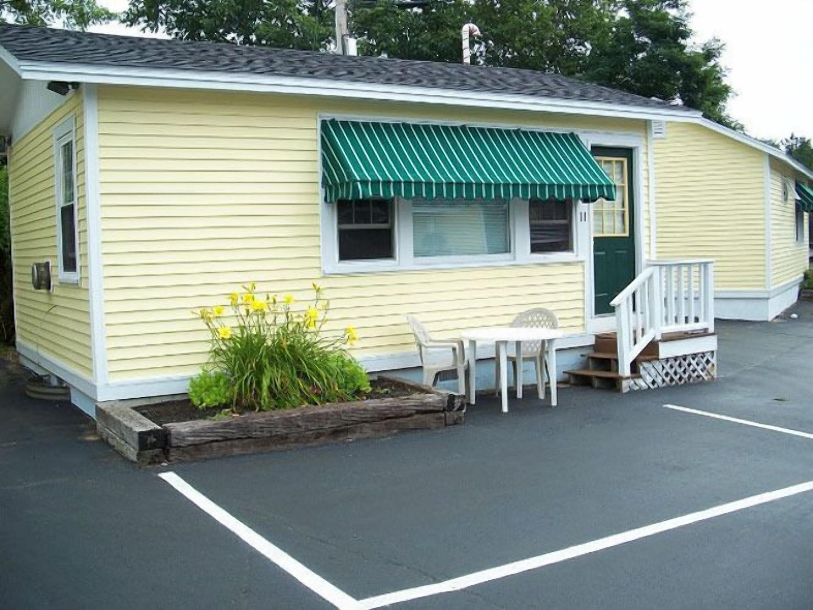 A small, yellow building with a green awning, a porch with white railings, and a small garden with yellow flowers.