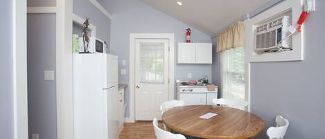 Cocina compacta con muebles blancos, microondas y estufa. Comedor con mesa de madera y sillas blancas.