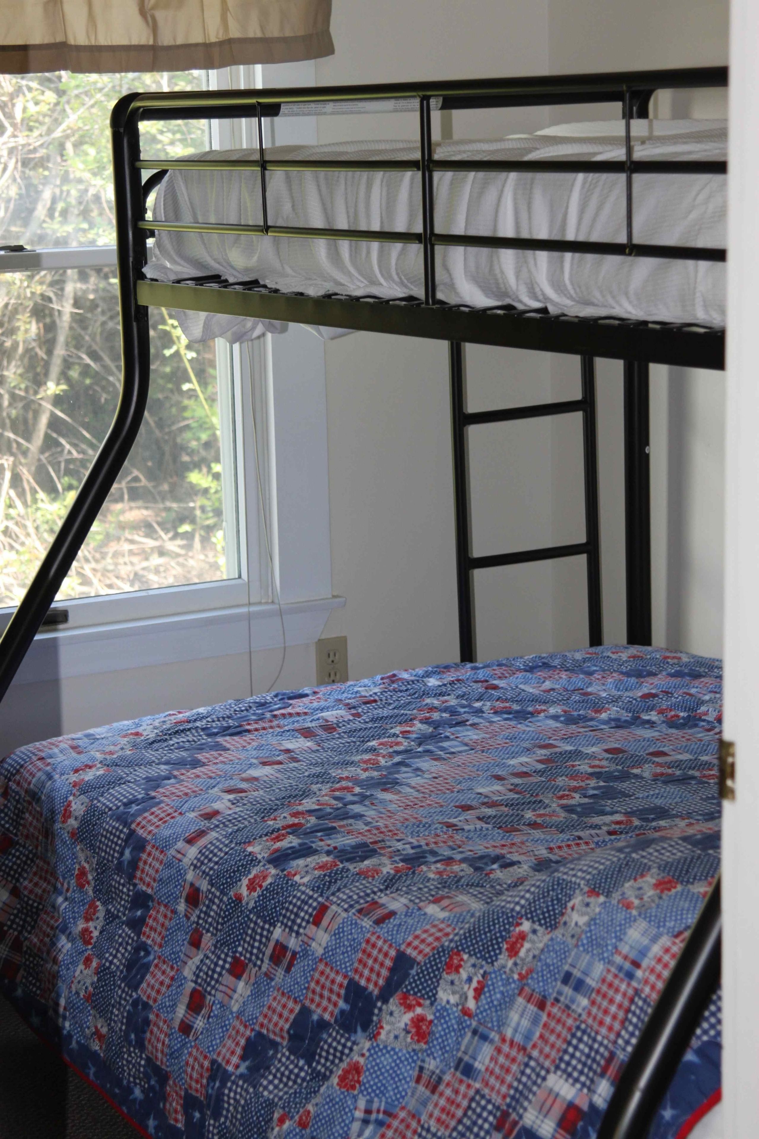 Un lit superposé avec une couvre-lit matelassé, une fenêtre donnant sur les arbres et une prise électrique au mur.
