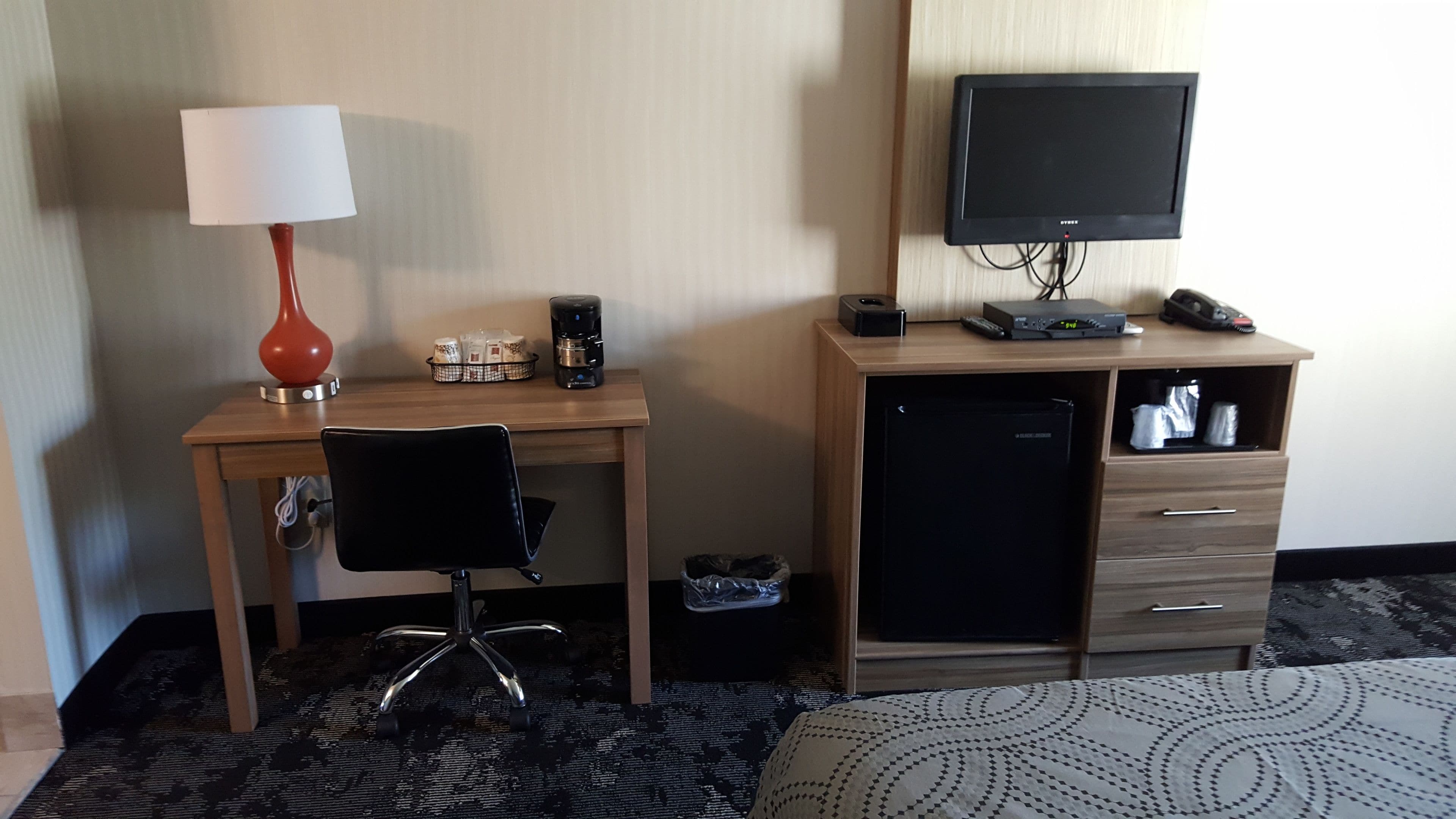 Ett hotellrum med ett skrivbord i trä, en svart kontorsstol, en tv på ett stativ, ett litet kylskåp och ett nattduksbord med en lampa.