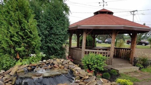 Un gazebo con tetto rosso e veranda in legno, circondato da un giardino con una piccola cascata e diverse piante.