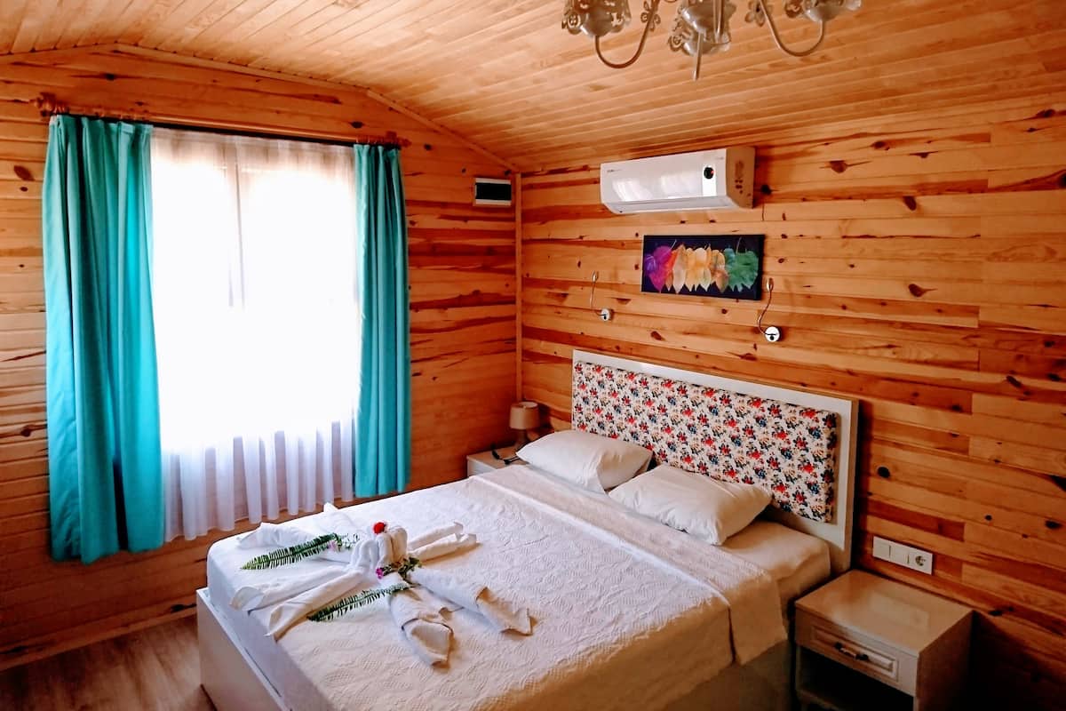 A wooden cabin-style room with a bed, a nightstand, a wall-mounted air conditioner, and a window with curtains.
