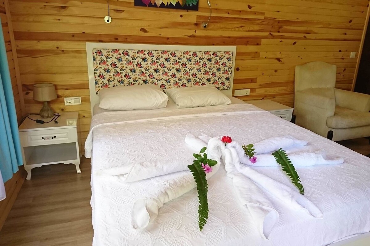 A wooden-paneled room with a bed, a chair, a nightstand, and a wall-mounted lamp.
