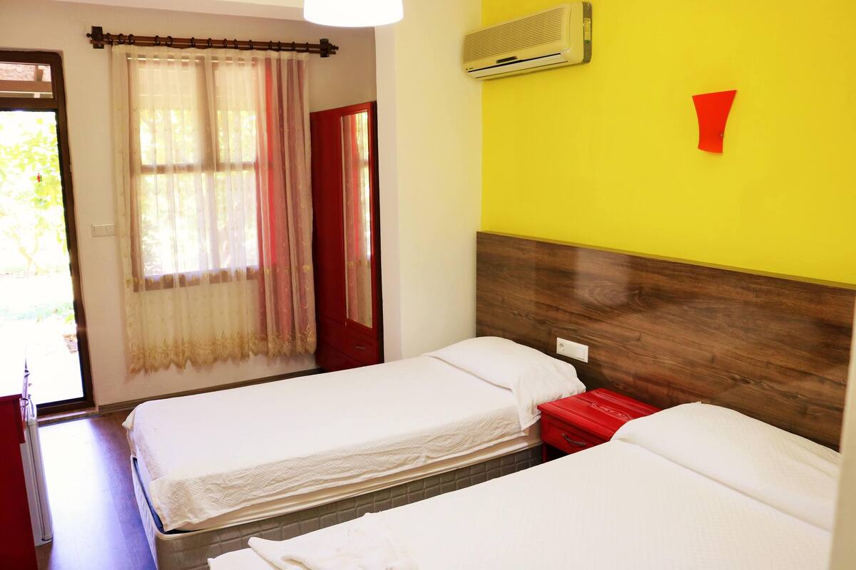 A hotel room with two single beds, a wooden headboard, a red accent pillow, a window with sheer curtains, and an air conditioning unit on the wall.