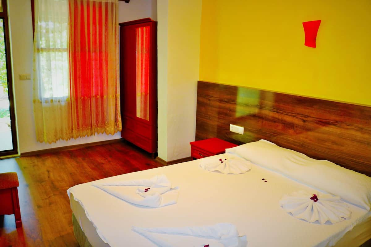 A hotel room with a wooden floor, a bed with white linens and towels, a red door, and a yellow wall with a red number sign.