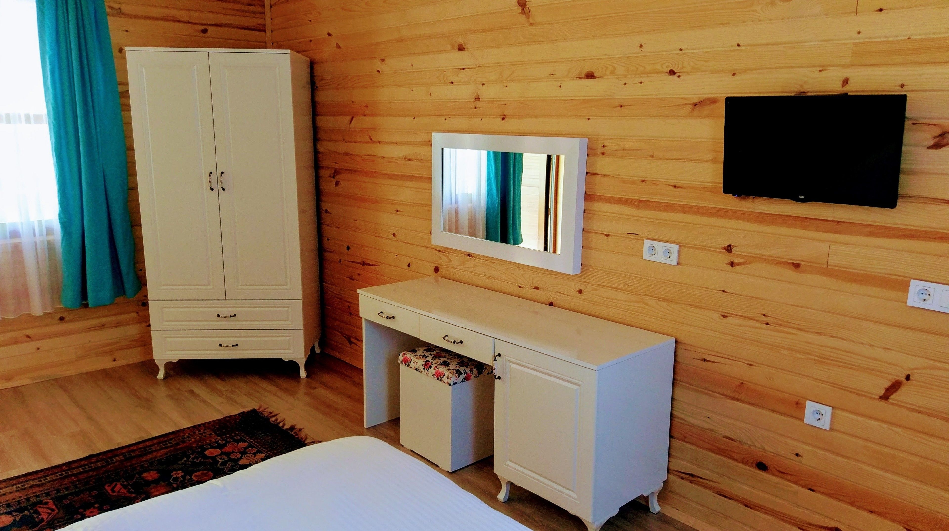 A room with wooden walls, a white wardrobe, a small desk with drawers, a mirror, and a bed.
