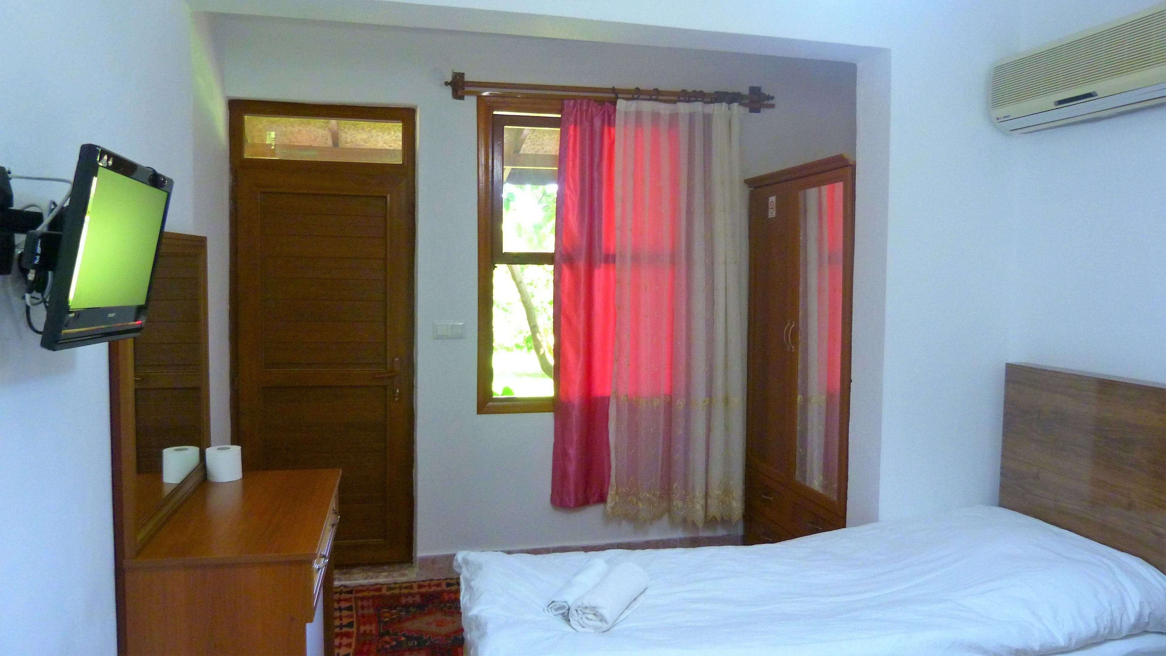 A hotel room with a bed, a desk with a computer monitor, a window with curtains, and a door.