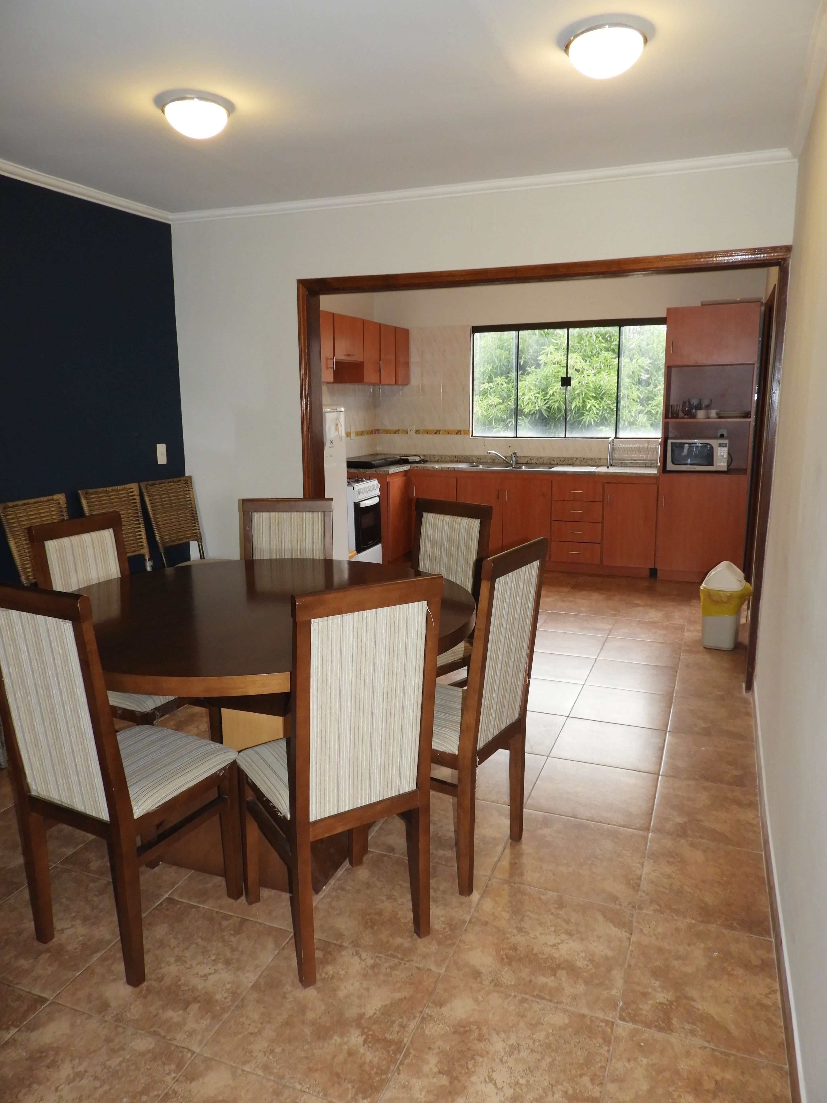 A dining area with a wooden table and chairs, a kitchen with wooden cabinets and a window, and a trash bin.