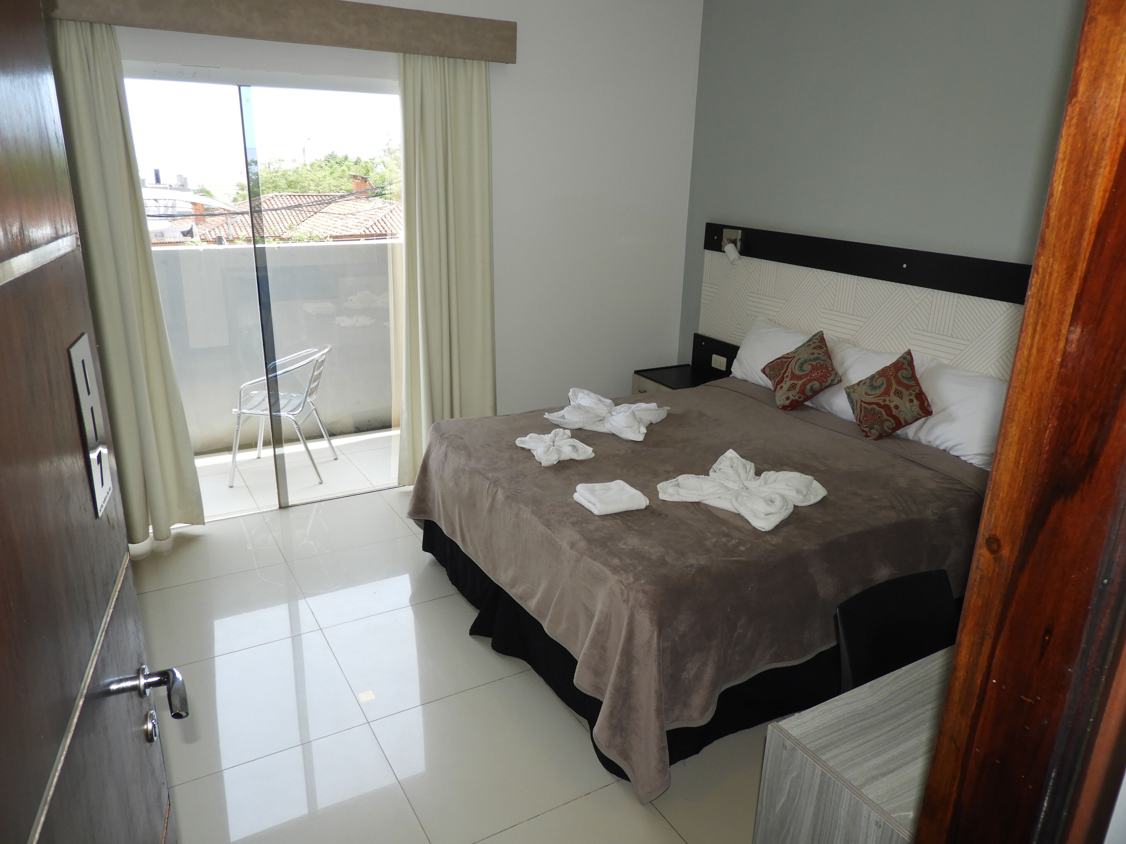 Habitación de hotel con cama, mesita de noche, una silla, balcón con vistas y puerta con manija.
