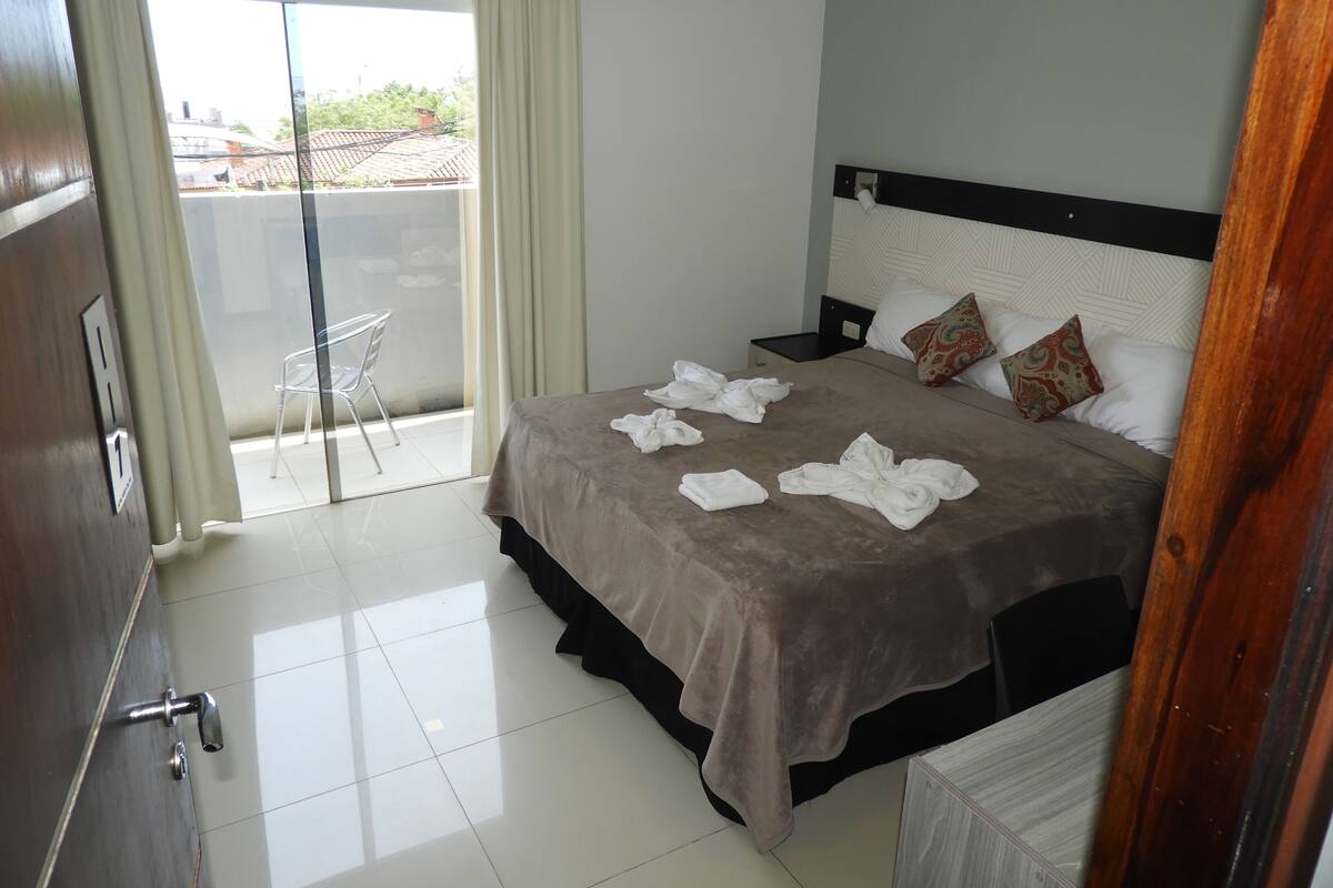 A hotel room with a bed, a nightstand, a chair, a balcony with a view, and a door with a handle.