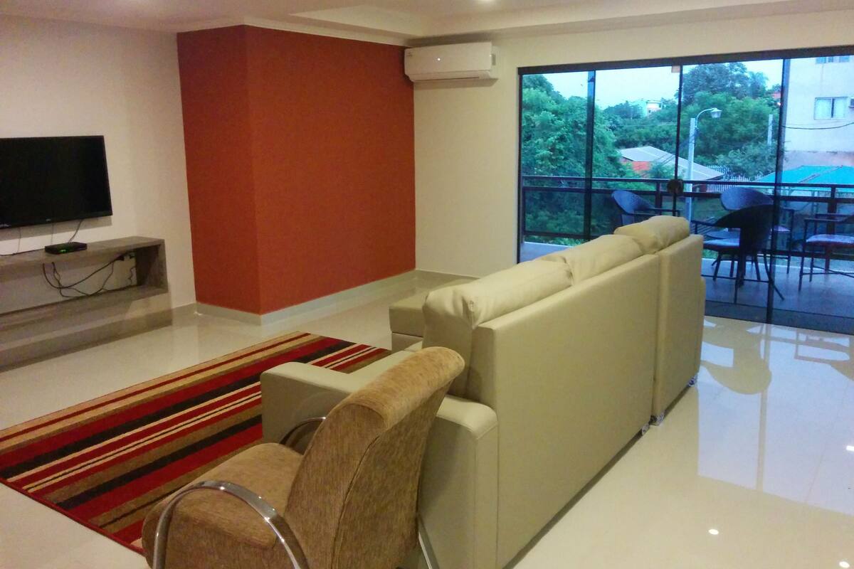 A modern living room with a sofa, a flat-screen TV, and a large window offering a view of the outdoors.